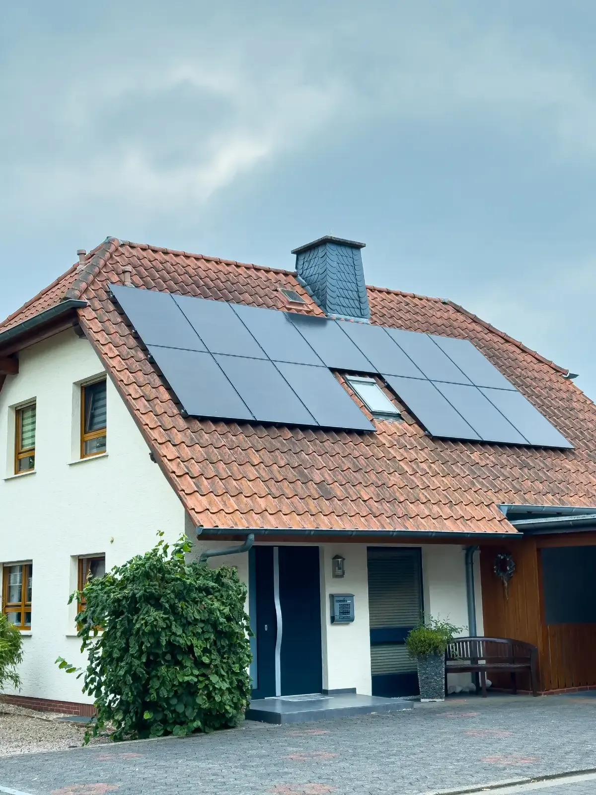 A two-storey rendered house with a terracotta tile roof carrying two rows of black solar panels arranged around two skylights, under an overcast sky.